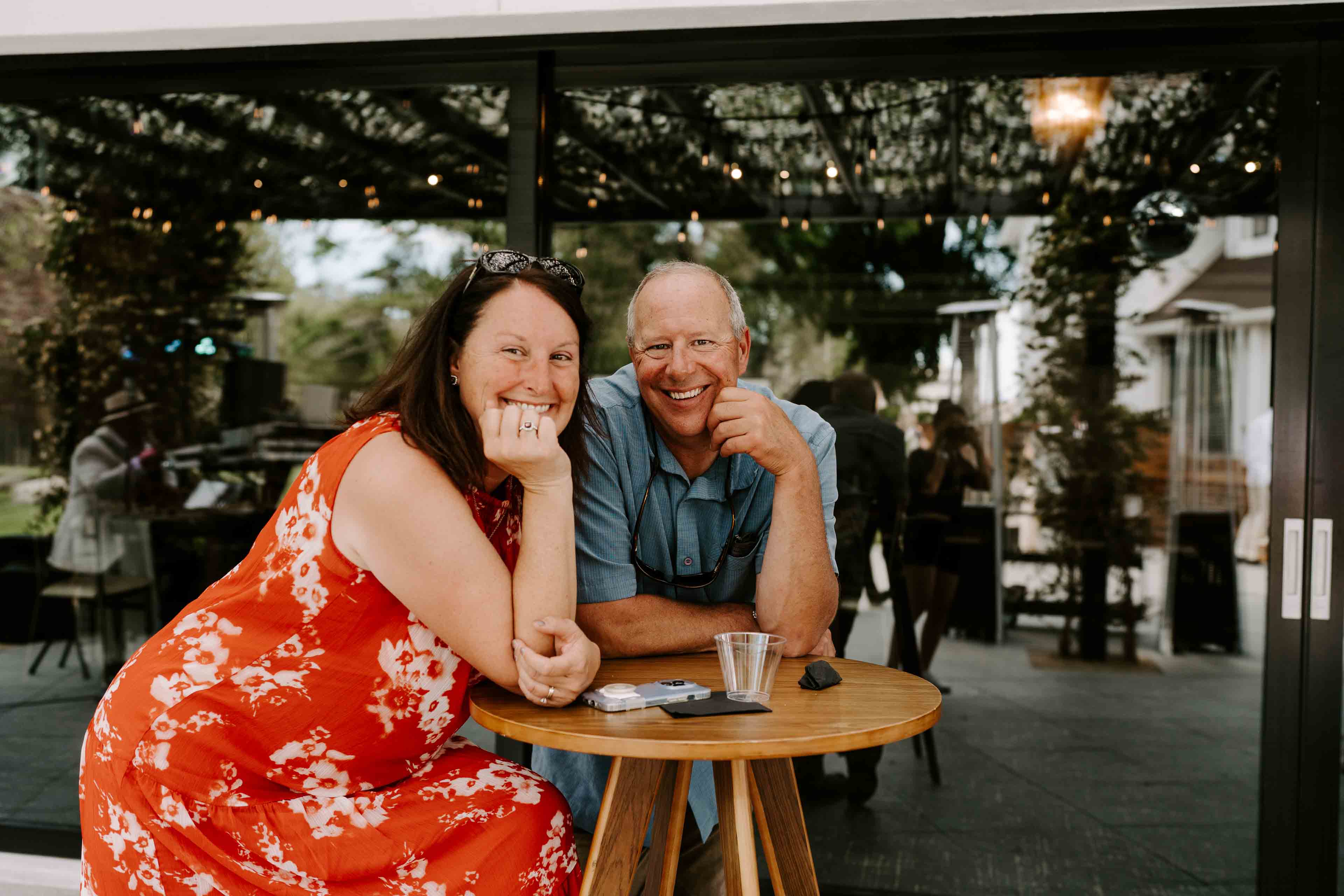 The Elm Estate A couple stting at an outside table with drinks smiling at the camera