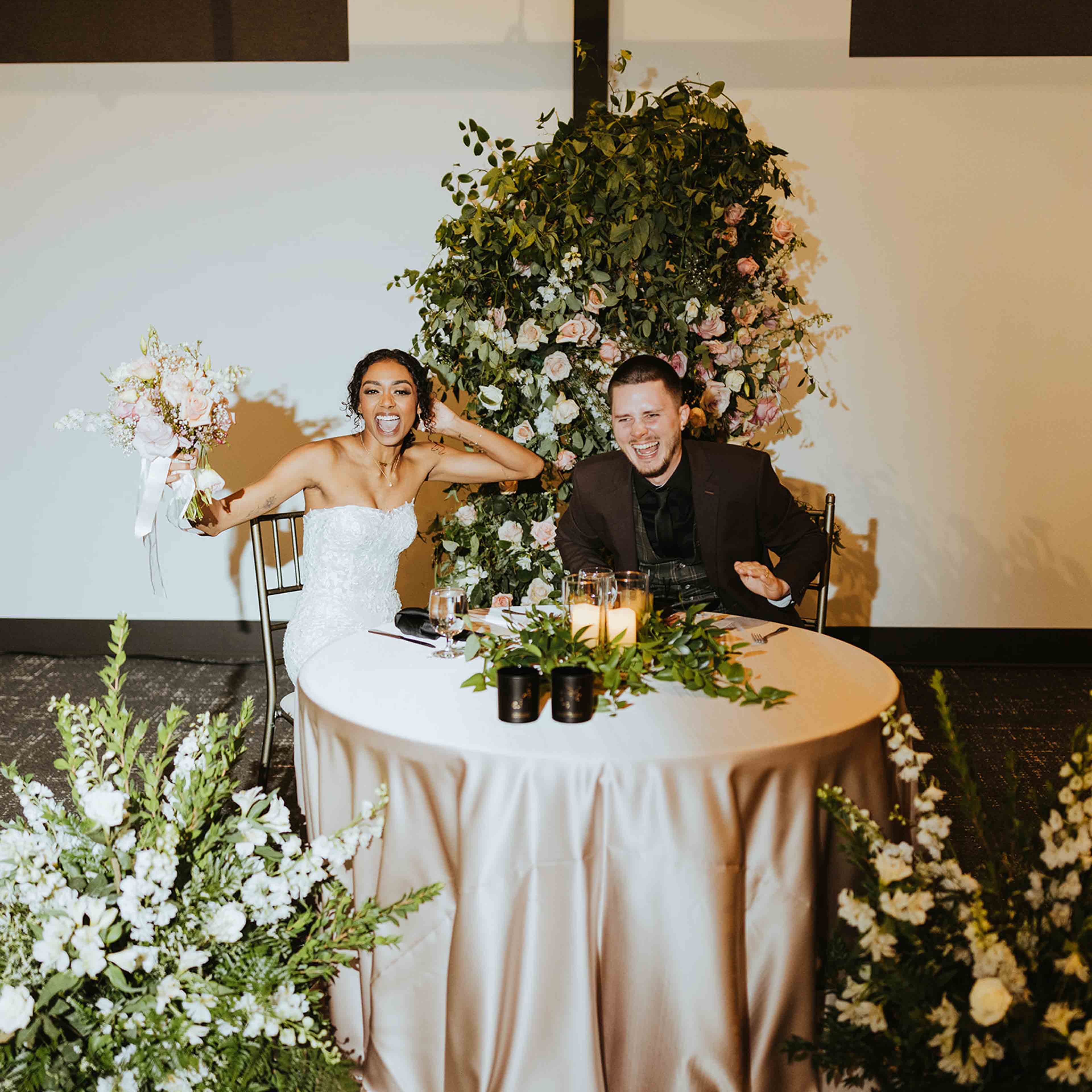 The Elm Estate Bride and goom sitting at a round table with big floral arrangements and smiling at the camera