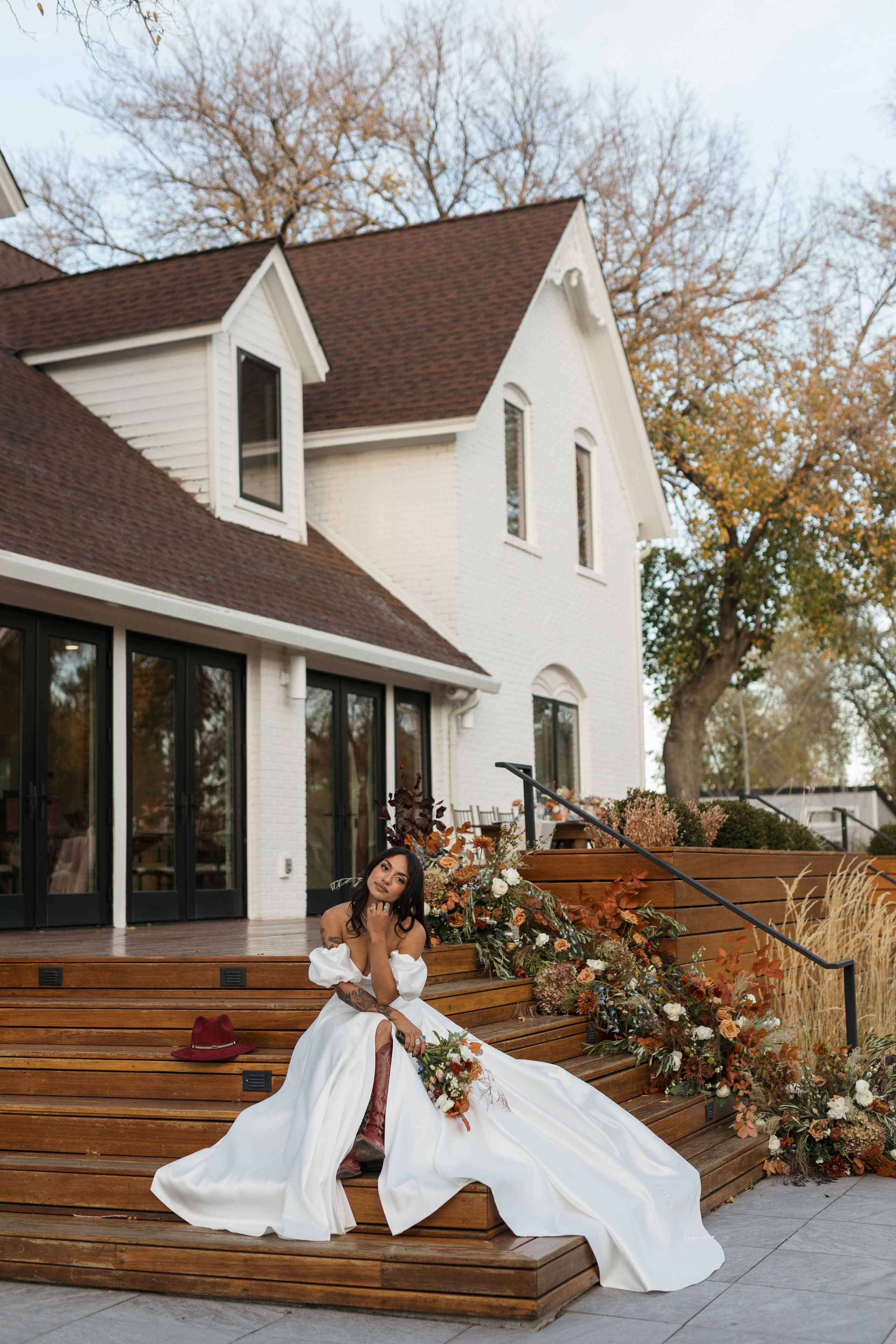 a bride sitting on the steps of a wooden deck outside a white building surrounded by fall colored floral arrangements