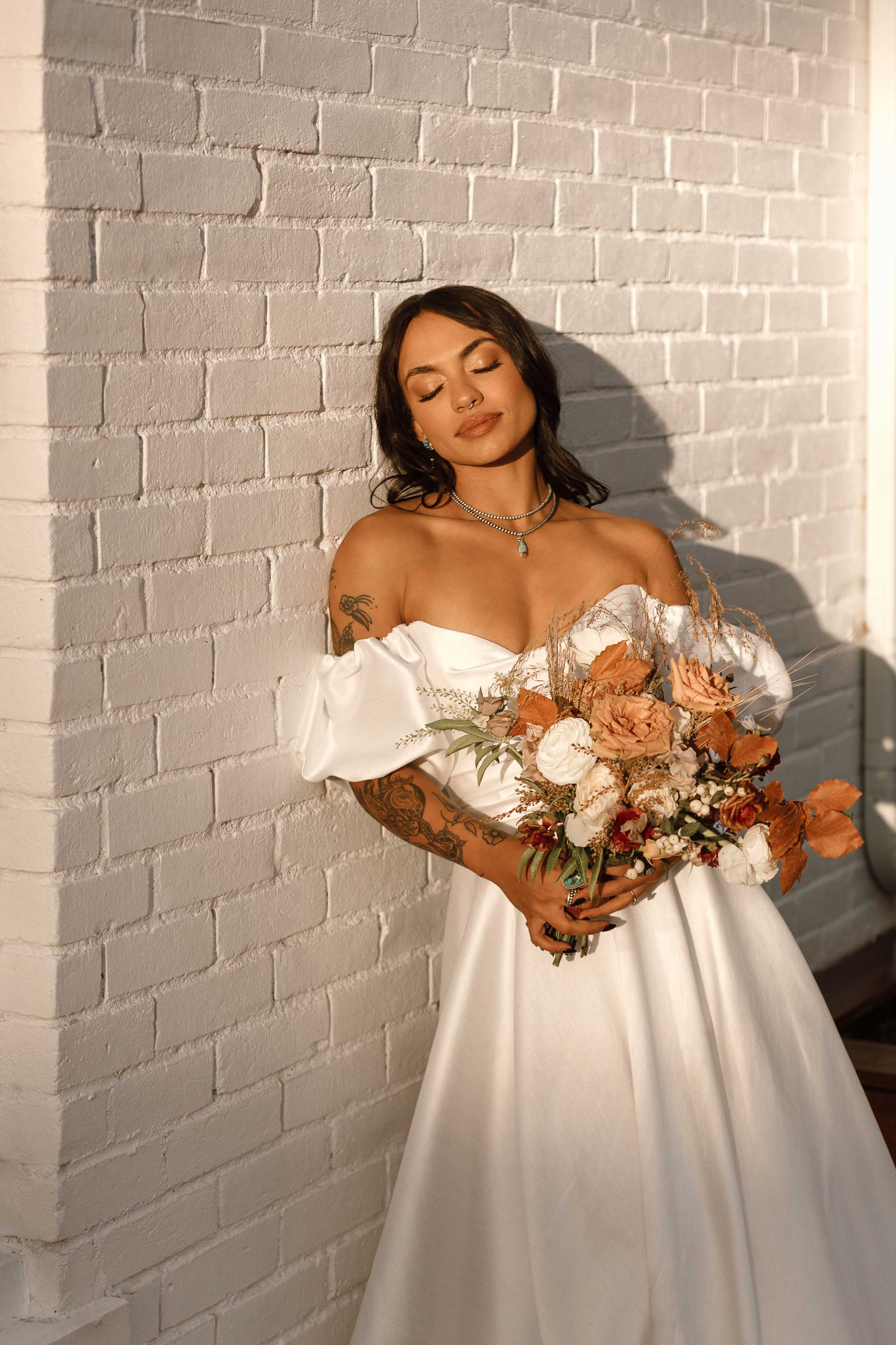 a bride standing against a white brick wall wearing a white gown and holding a bouquet
